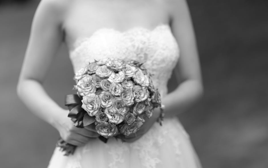A bride in a strapless lace wedding gown is holding a round bridal bouquet composed of tightly clustered, pale roses with ruffled petals, wrapped with a dark satin ribbon. The bouquet's flowers appear