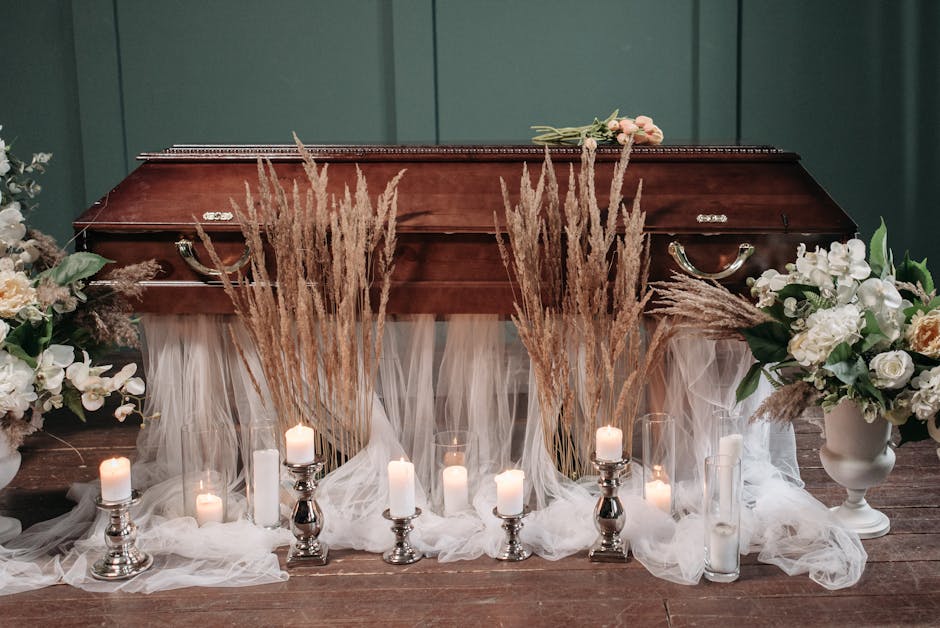A wooden casket is decorated with arrangements of white and cream roses, lilies, and greenery, flanked by bouquets in white urns, set against a dark green wall. Tall dried pampas grass and delicate wh