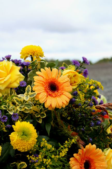A vibrant floral arrangement comprising large yellow roses, orange gerbera daisies with dark centers, and clusters of small purple and yellow flowers. The bouquet features lush green foliage and varie