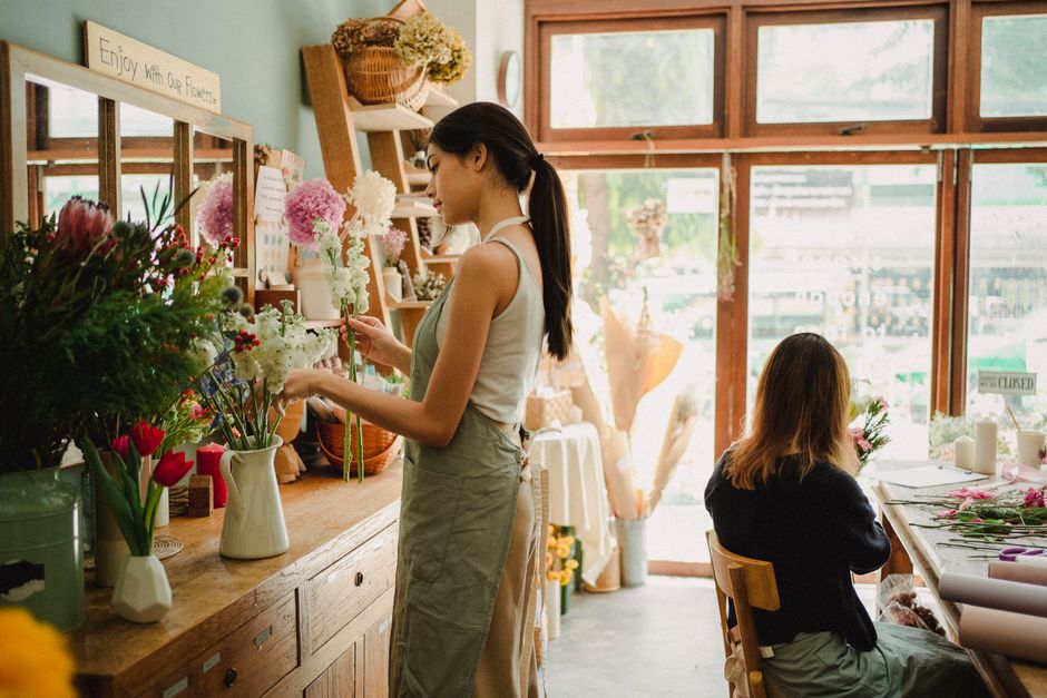 A young woman in a white sleeveless top and beige apron stands in a bright florist shop, carefully arranging a bouquet of pink and white hydrangeas, white tulips, and greenery on a wooden counter. The