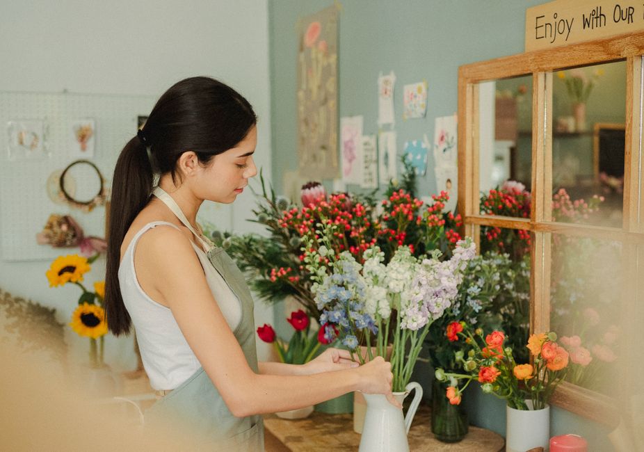 A young woman with dark brown hair tied in a low ponytail, wearing a light-colored sleeveless top and a beige apron, arranging a bouquet of fresh, pastel-colored flowers in a white ceramic pitcher ins