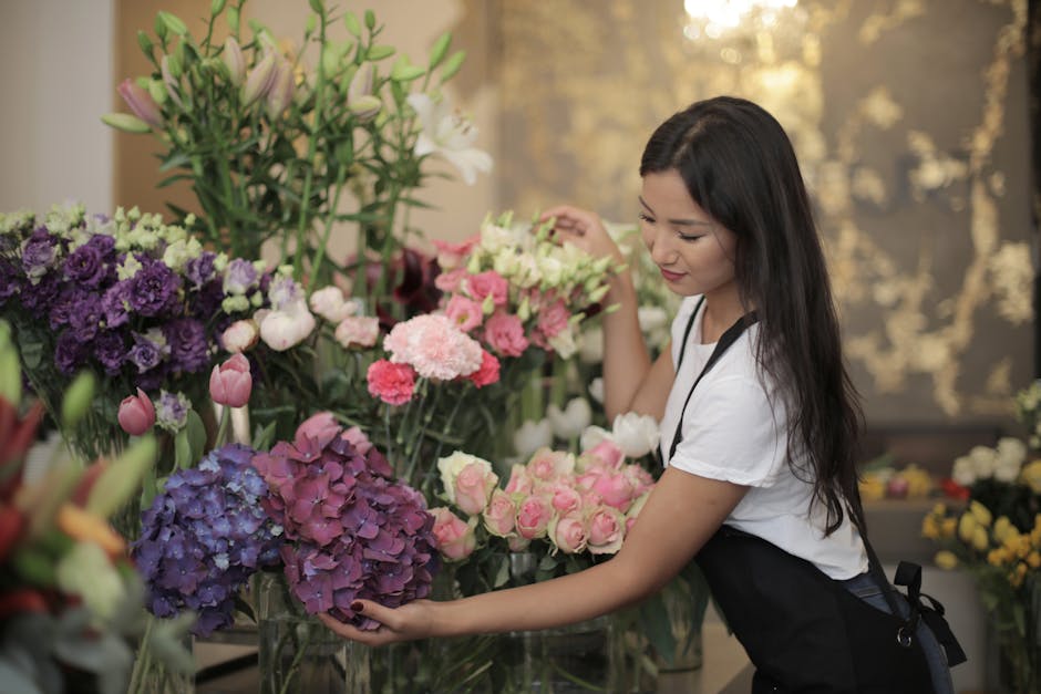 A young female florist with long dark hair arranging a bouquet of fresh flowers in a well-lit floral shop. She is wearing a white t-shirt and a black apron, and is carefully handling a cluster of pink