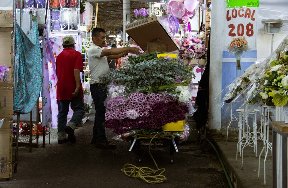 A florist at Greenwich Market is arranging a vibrant bouquet of flowers, including pink and purple chrysanthemums, and white blossoms, on a yellow table. The person is holding a cardboard box, possibl