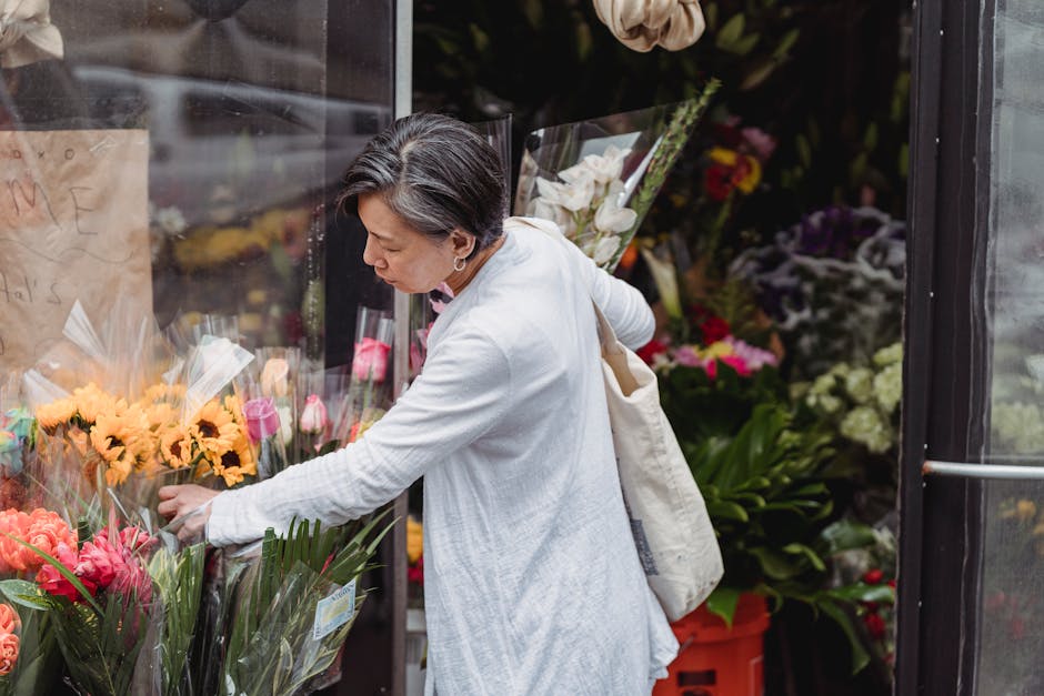 A woman with short gray hair, wearing a white long-sleeve shirt and carrying a cream-colored tote bag on her shoulder, selecting fresh flowers at an outdoor flower shop. She is reaching out to pick a