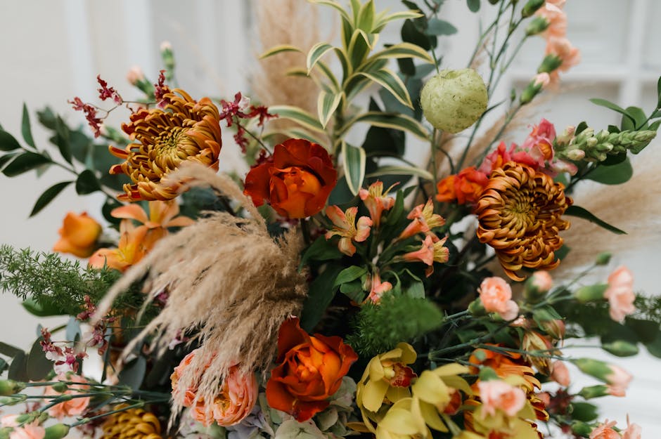 A vibrant floral arrangement featuring orange roses, large chrysanthemum blooms with curled petals, and delicate pink and yellow blossoms, all intricately arranged with green foliage and variegated le